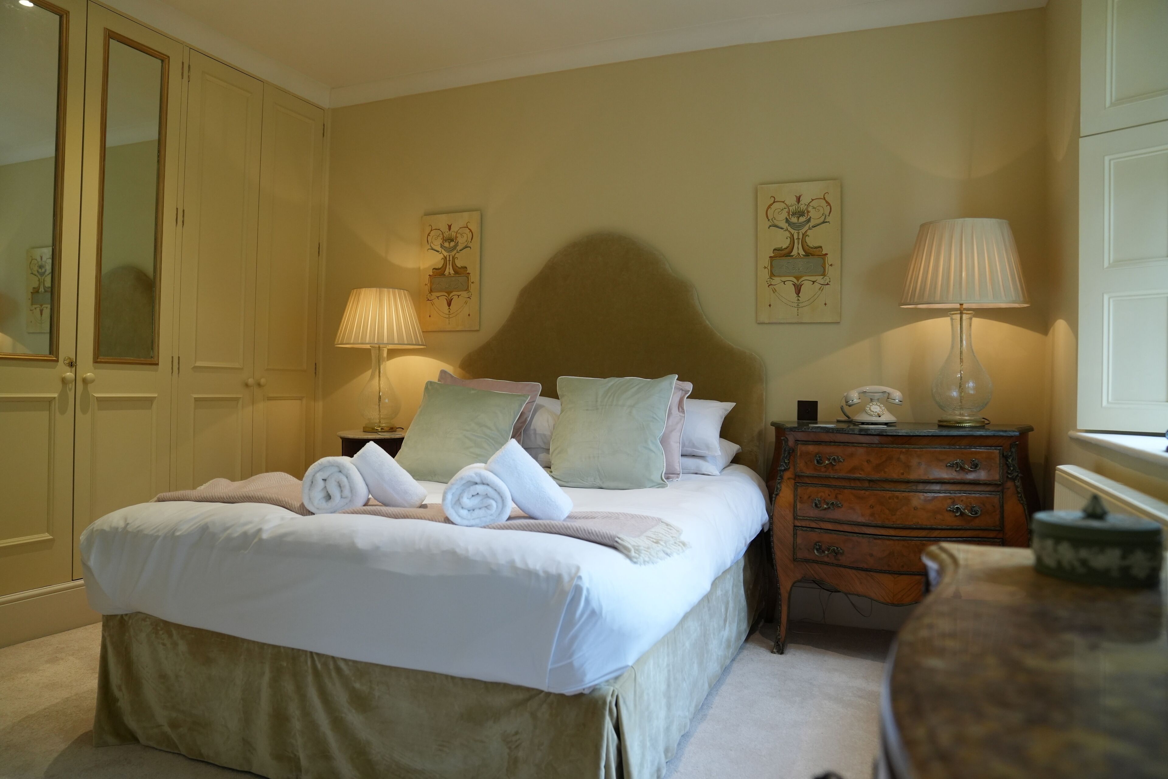 A neatly made bed with pillows and rolled towels, a wooden nightstand with a lamp, a dresser, and a mirror.