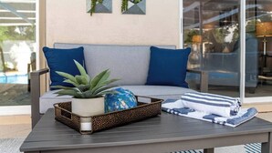 Ein moderner Außenbereich mit einer grauen Couch, blauen Kissen, einer Zierwand und einem Tisch mit Pflanze und blauer Vase.