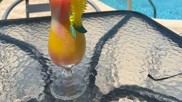Un vaso con una bebida colorida y una pajita, sobre una mesa cerca de una piscina con tumbonas y un edificio al fondo.