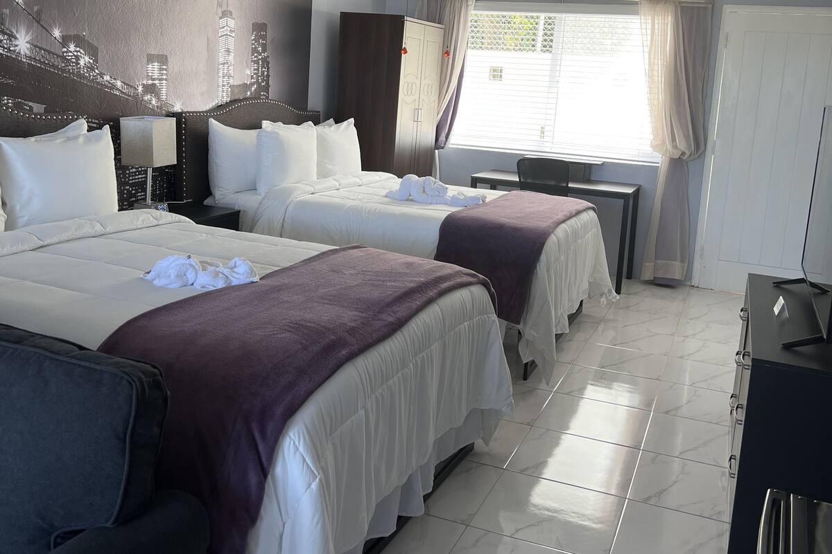 A hotel room with two beds, a ceiling fan, an air conditioning unit, a desk with a chair, and a window with curtains.