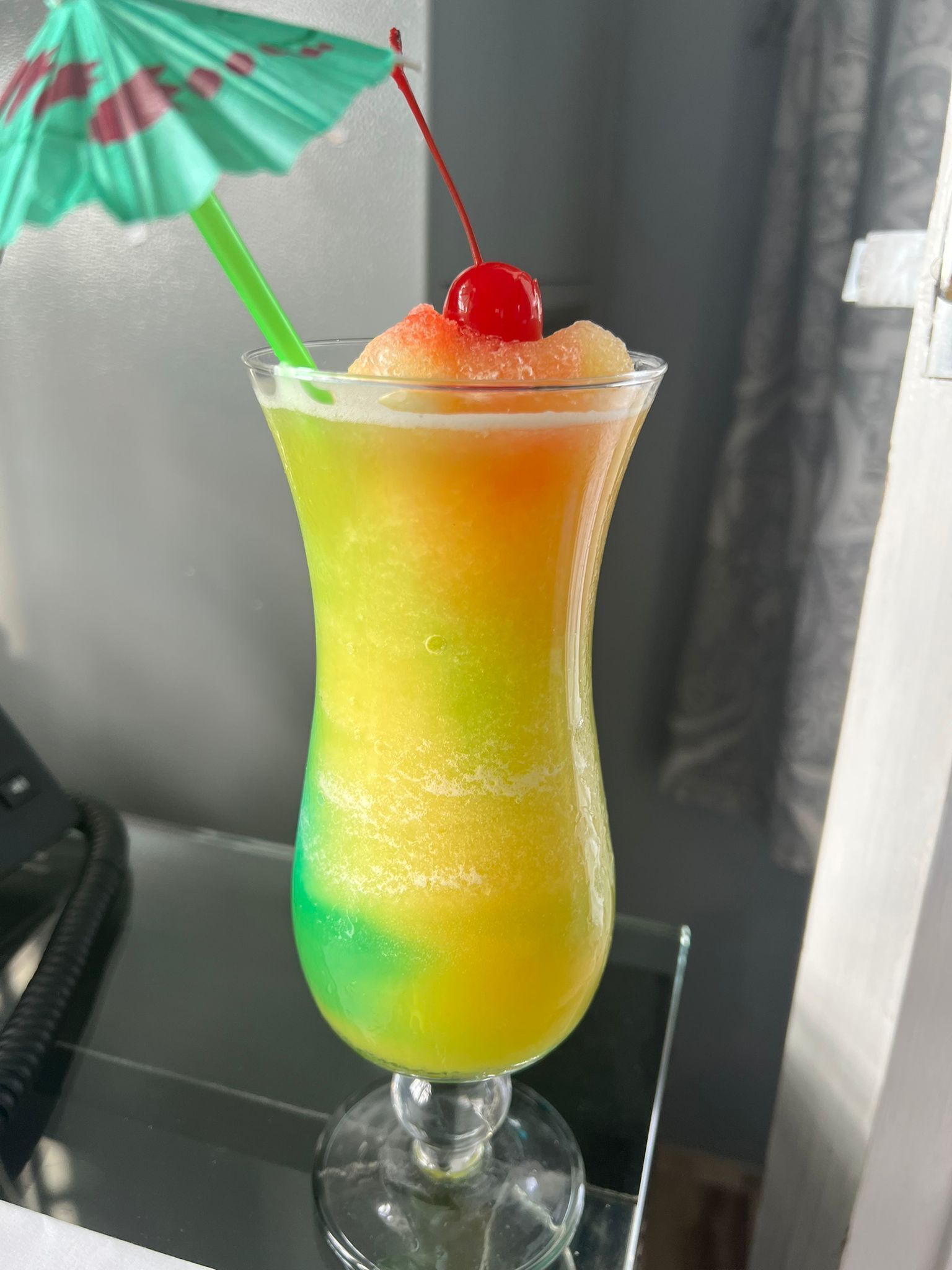 A cocktail in a tall glass with a gradient color from green to yellow, topped with a cherry.