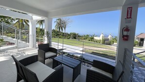Un área de estar al aire libre techada con mobiliario de mimbre y una mesa de vidrio, con vistas a un jardín ajardinado y al océano.