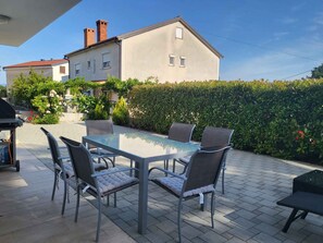 Une terrasse extérieure avec une table et des chaises en verre, un barbecue et une haie.