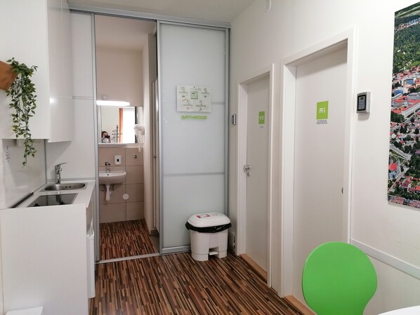 A narrow hallway with a wooden floor, a sink, and a toilet. There are signs indicating the bathroom and a green chair.