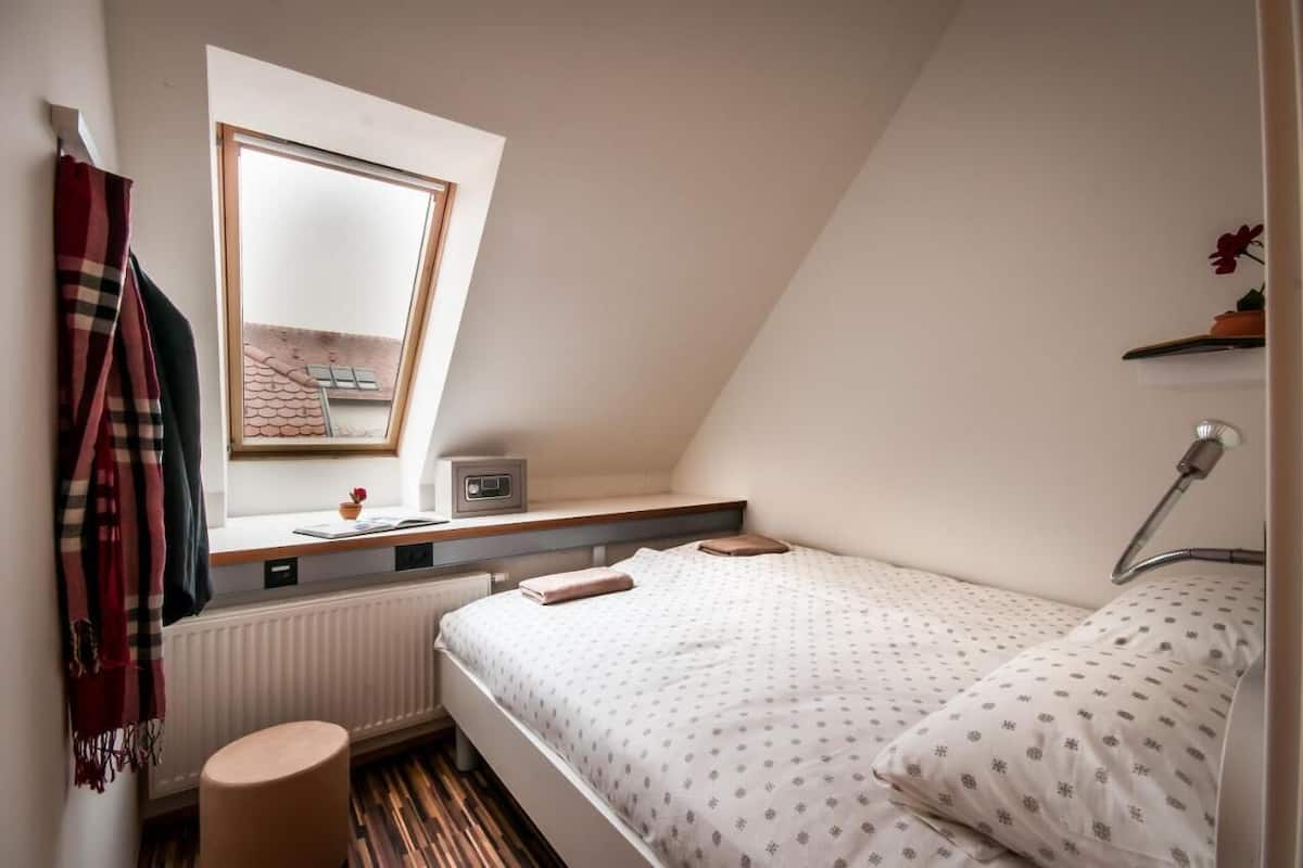 Ein kleines, gemütliches Schlafzimmer mit schrägen Wänden, einem Fenster, einem Nachttisch mit Radio, einer Lampe und einem Bett mit gemusterten Betttextilien.