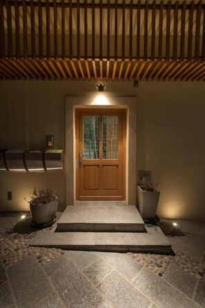 A wooden door with a glass panel, flanked by two potted plants, leading to a stone-paved entrance.