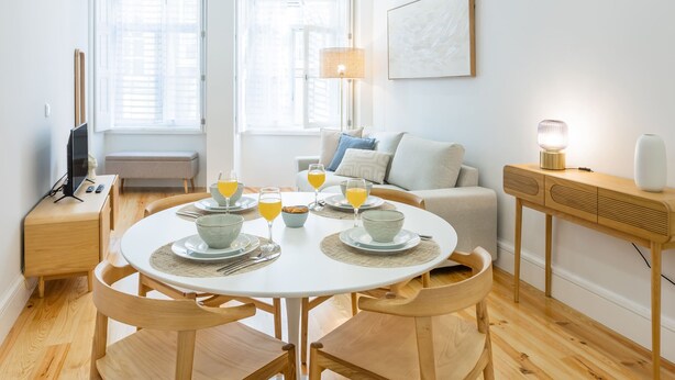 Ein moderner Essbereich mit einem runden Tisch, vier Holzstühlen und einer Bank. Links befindet sich ein Fernseher auf einem Schrank und auf einem Beistelltisch eine Lampe.