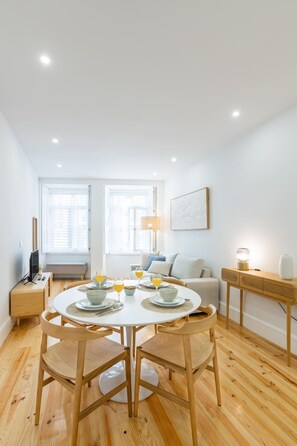 A modern dining area with a round table, four wooden chairs, and a bench. There is a television on a stand to the left and a lamp on a side table. - Herança dos Mercadores (Porto)