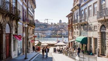 A cobblestone street with buildings on either side, a river in the background, and outdoor seating areas.