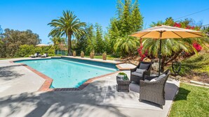 Une piscine dont le bord est revêtu de carreaux, entourée d’une terrasse aménagée avec des meubles en osier, une table avec des boissons et un grand parasol.