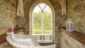 A cozy room with a stone wall, a large arched window, a wicker chair, and a white lantern on a shelf. - The Wagon House (Launceston)