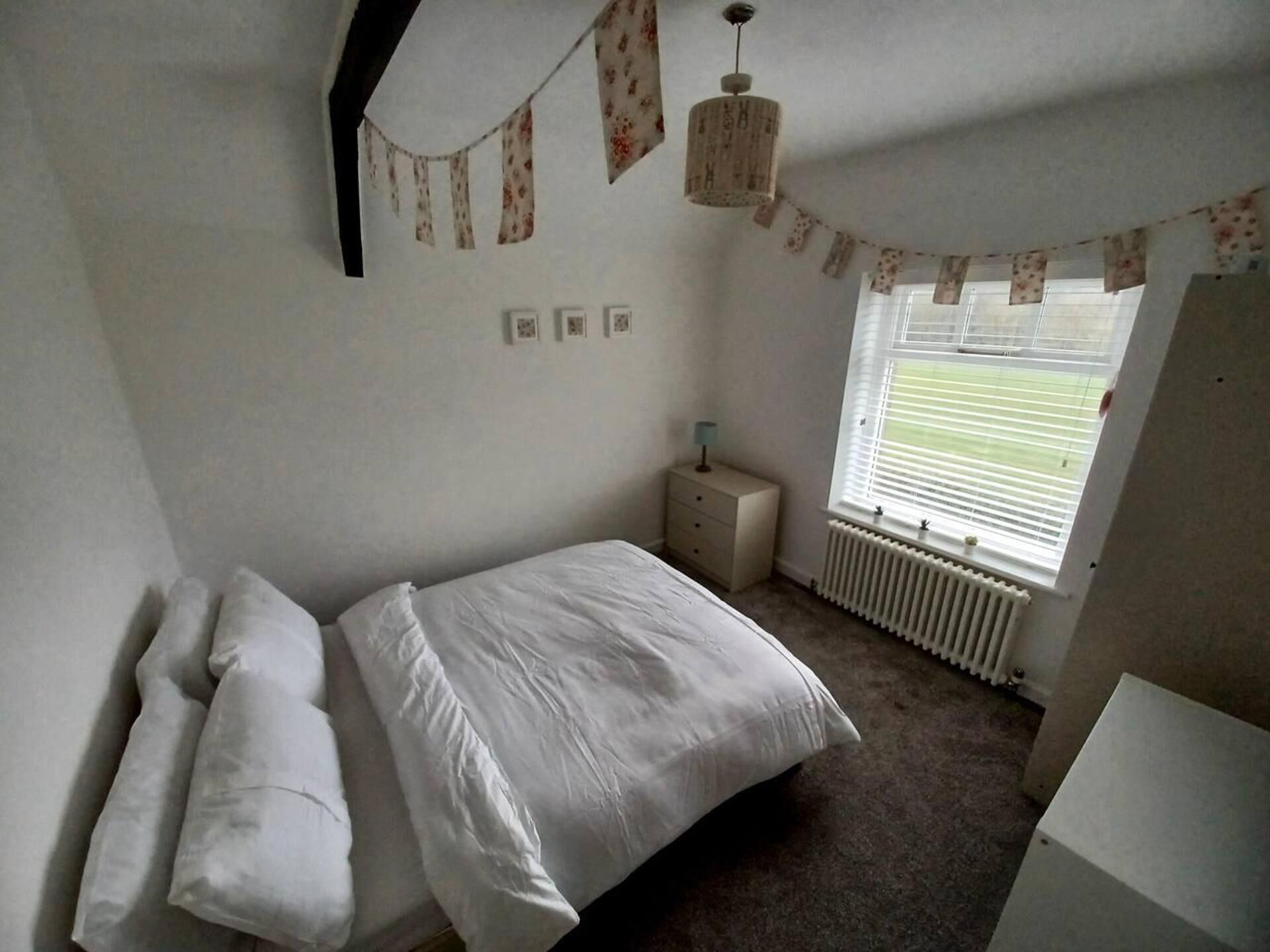 A bedroom with a bed, a nightstand, a lamp, a window with blinds, and a radiator.