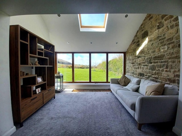 A living room with a stone wall, a large window with a view of a green field, a grey sofa, and a wooden bookshelf. - 3 Bed Cottage-sleeps 6-country Views-pet Friendly (Sunderland)