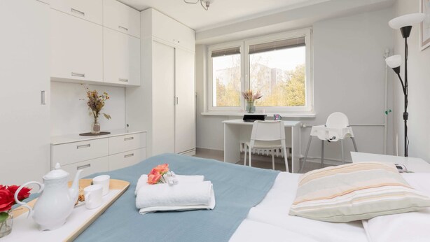 A modern hotel room with a bed, a desk, a chair, and a window with a view of buildings. - Spacious Apartment | 2 Bedrooms, Balcony, Close to Metro (Warszawa)