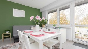 A dining area with a white table set for four, a green wall, and a view through large windows. - Spacious Apartment | 2 Bedrooms, Balcony, Close to Metro (Warszawa)