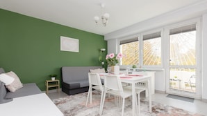 A modern living room with a green accent wall, a grey sofa, a white dining table with chairs, and a large window with a view of a tree. - Spacious Apartment | 2 Bedrooms, Balcony, Close to Metro (Warszawa)