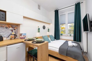 A compact hotel room with a kitchenette, a bed with a grey checkered bedspread, teal pillows, and a window with curtains. - Stylish Studio in Krakow | Close to Main Station and Market Square (Kraków)