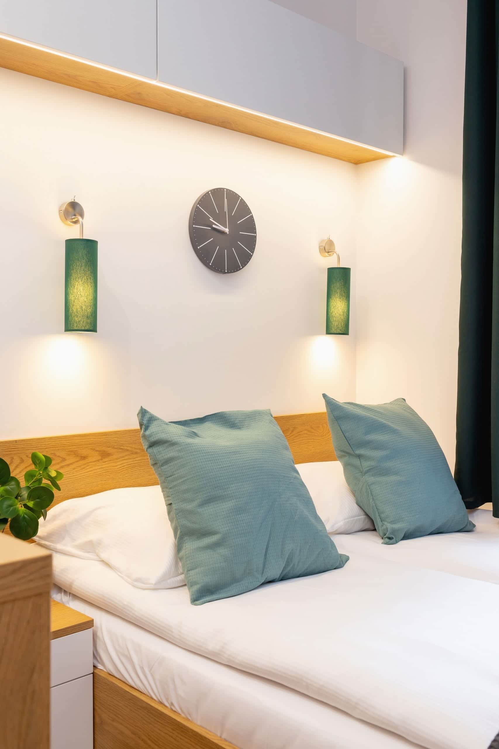 A neatly made bed with two pillows, a bedside table with a plant, and two wall-mounted clocks.