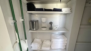 A small utility room with shelves holding cleaning supplies, a coffee maker, and folded towels. - Downtown Colorado Springs Studio (Colorado Springs)