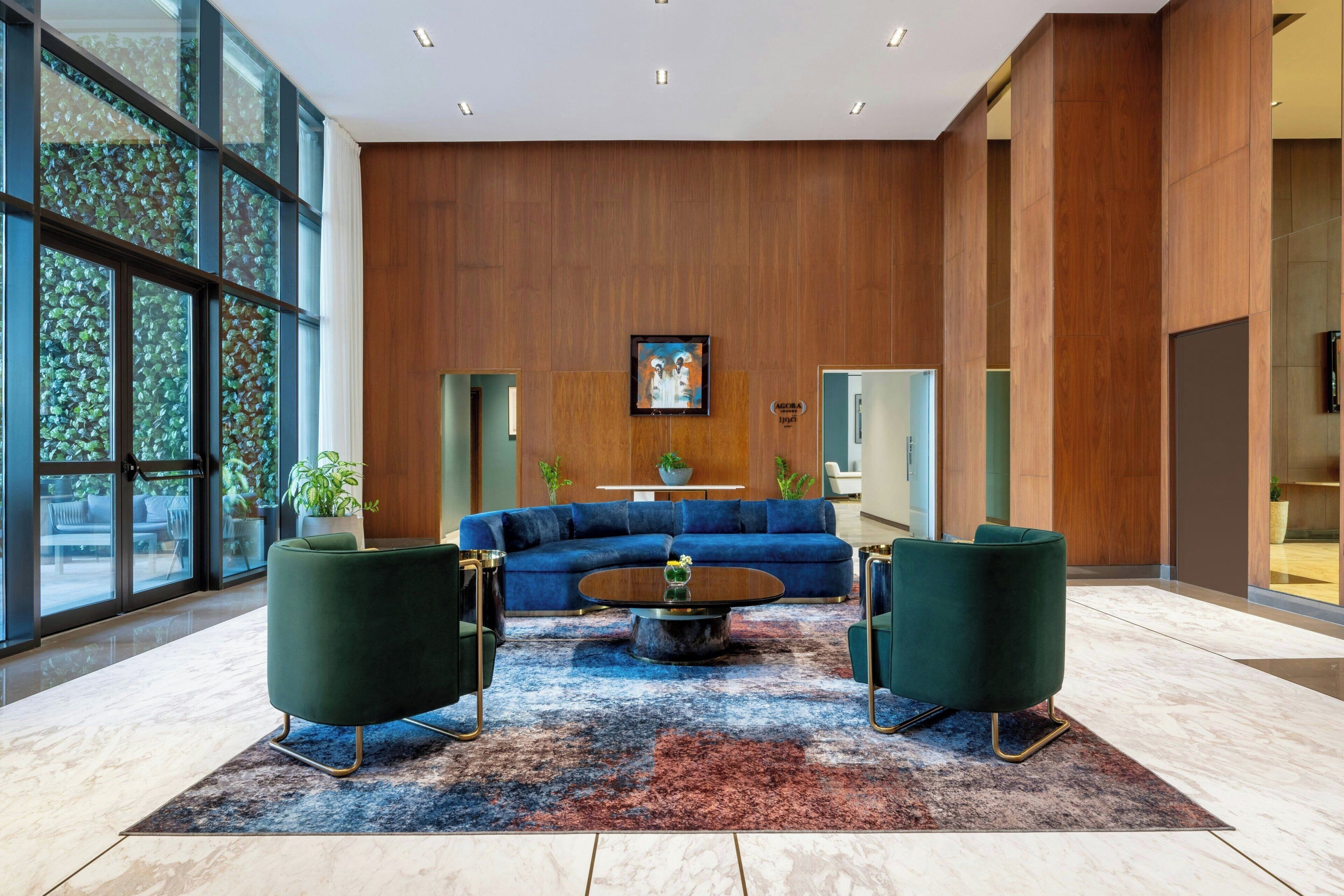 Ein modernes Wohnzimmer mit einem blauen Sofa, zwei grünen Sesseln, einem Couchtisch und einem großen Fenster mit Blick ins Grüne.