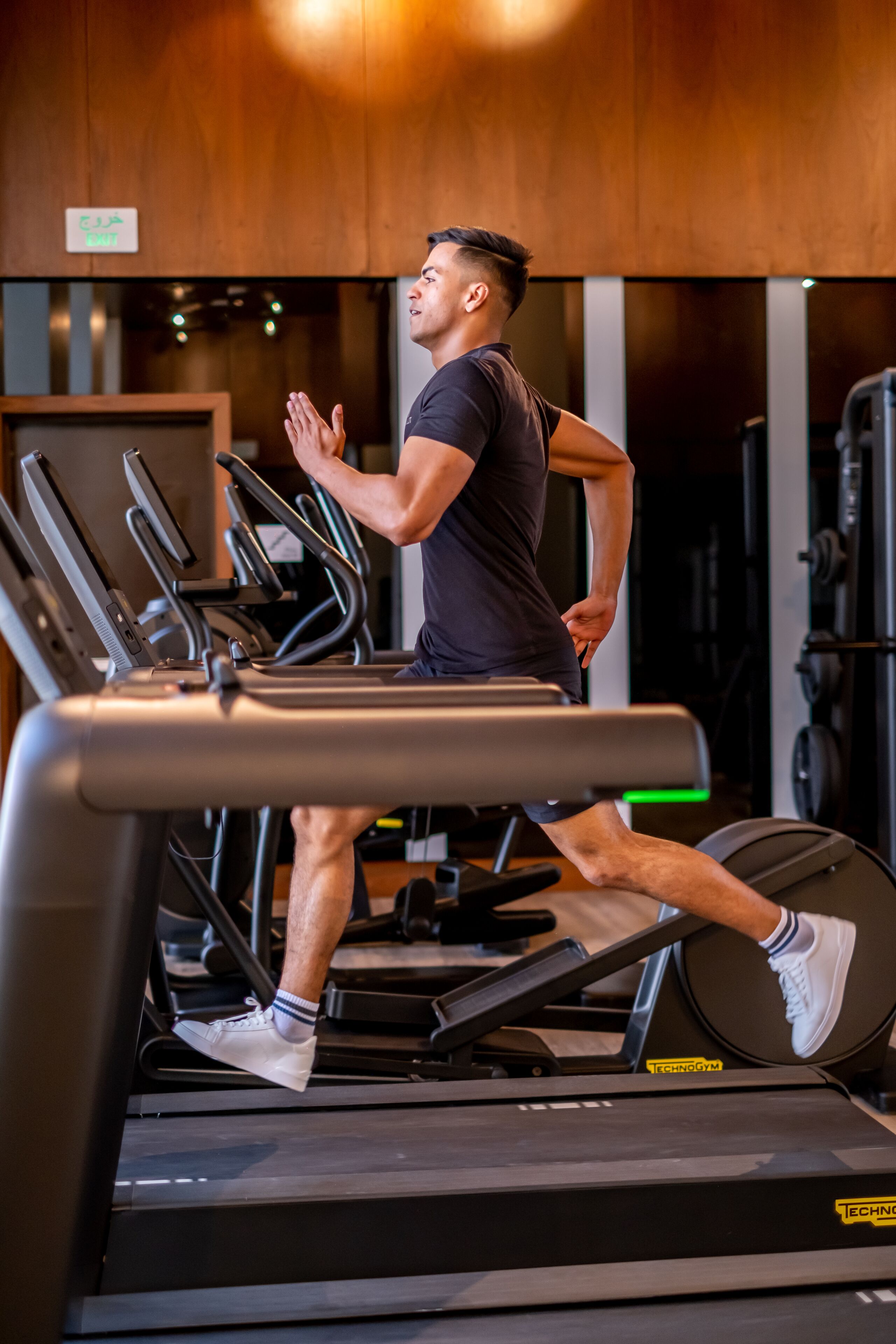 A person running on a treadmill in a gym with visible exercise equipment and a wooden wall in the background.