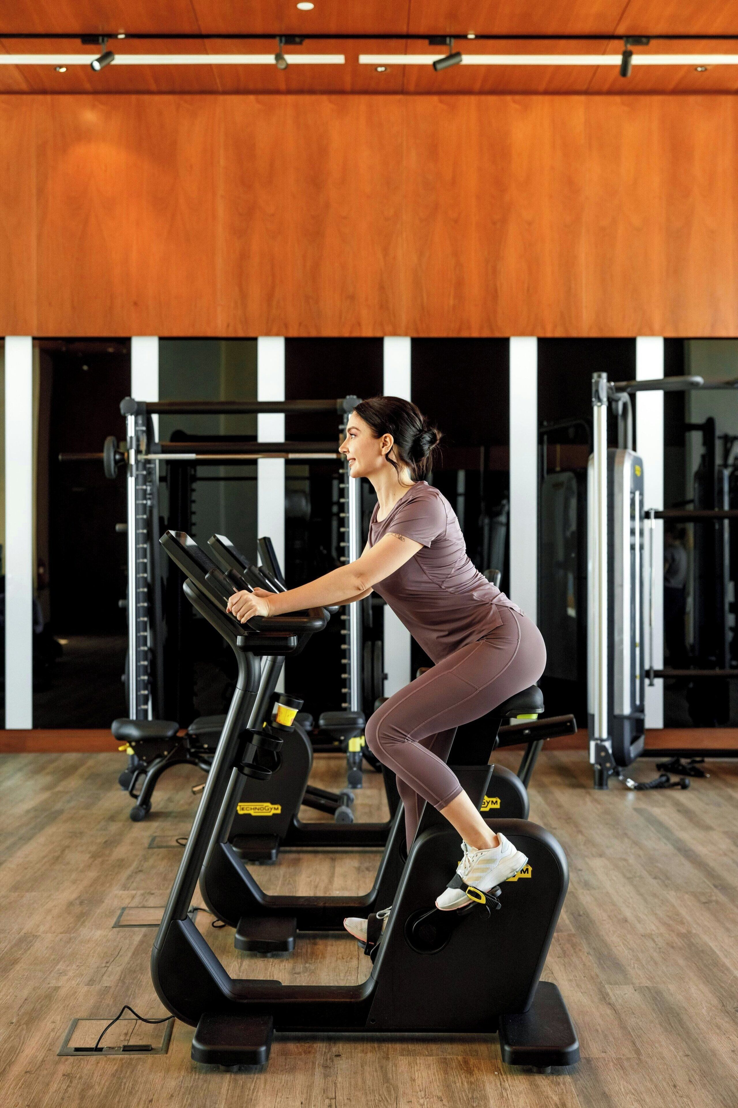 Un gimnasio con equipo de ejercicio, piso de madera y pared espejada.