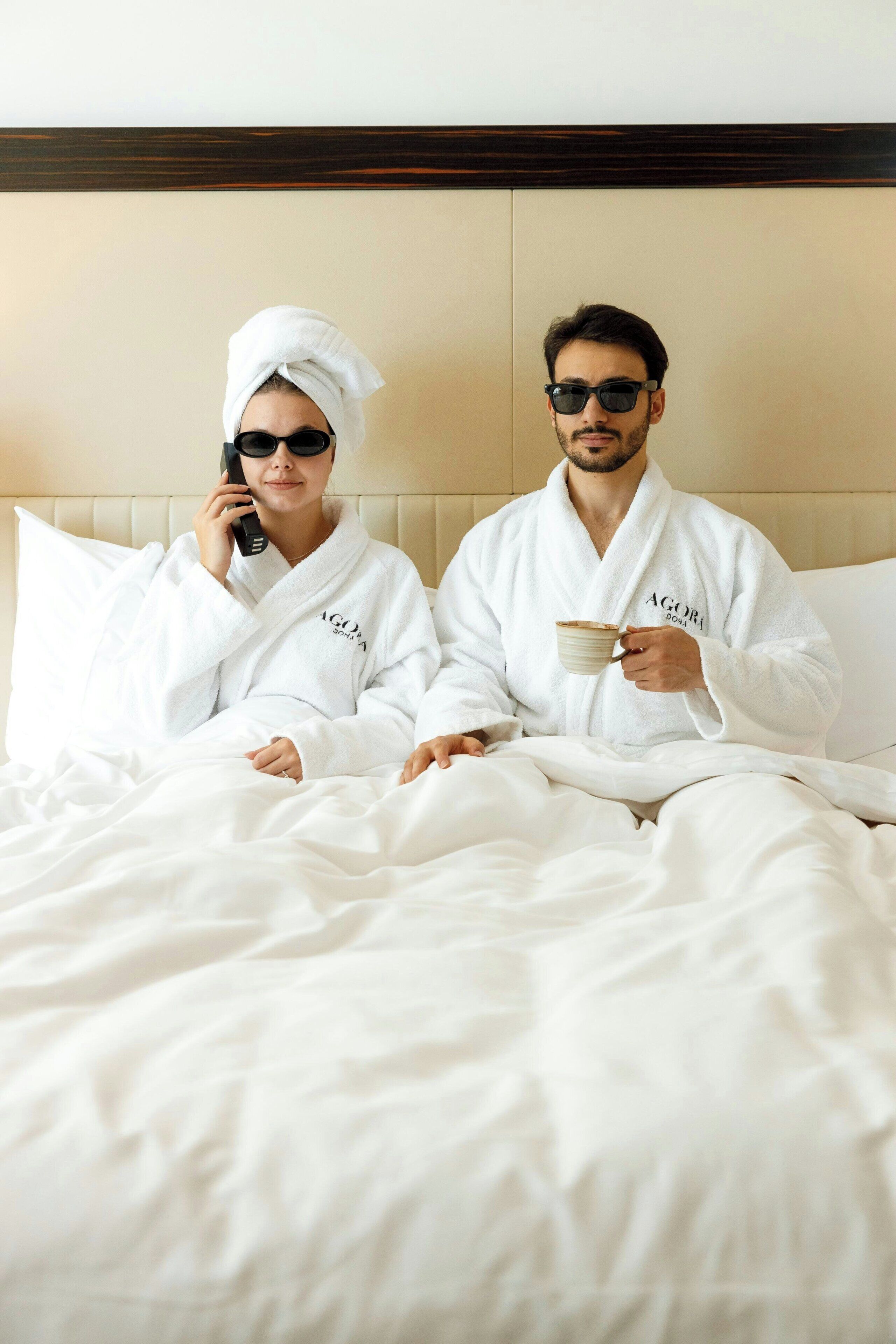 Dos personas con batas blancas sentadas en una cama con cabecera de madera, una sosteniendo una taza.