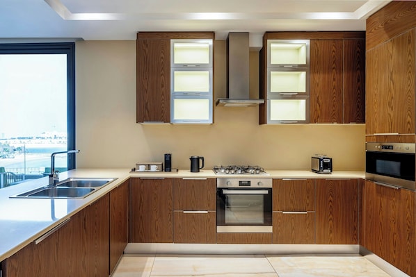 A modern kitchen with wooden cabinets, stainless steel appliances, and a large window providing a view of the city. - Agora, Doha, Autograph Collection (Doha)