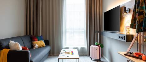 A modern hotel room with a sofa, a small table, a TV, a suitcase, and a desk with a lamp.