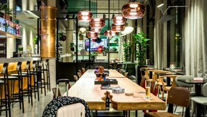 A modern dining area with a long wooden table, multiple chairs, and hanging copper pendant lights. - Moxy Dortmund (Dortmund)