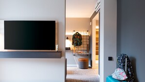 A modern hotel room with a flat-screen TV mounted on the wall, a bed with a white comforter, a suitcase, and a small round ottoman. - Moxy Dortmund (Dortmund)