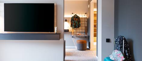 Una habitación de hotel moderna con un televisor de pantalla plana montado en la pared, una cama con colcha blanca, una maleta y un reposapiés redondo pequeño.