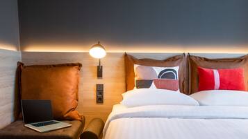 A hotel room with a bed, a laptop on a cushioned chair, a bedside lamp, and a wooden headboard.