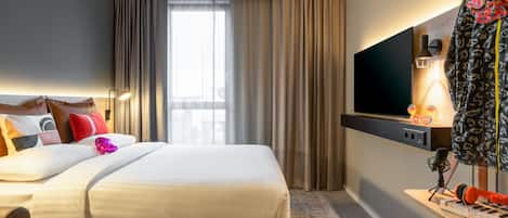 Una habitación de hotel con cama, televisión, escritorio con teclado y auriculares, y ventana con cortinas.