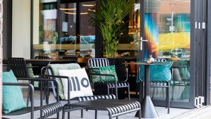 An outdoor seating area with metal chairs and tables, a large potted plant, and a glass door reflecting the interior. - Residence Inn by Marriott Dortmund (Dortmund)