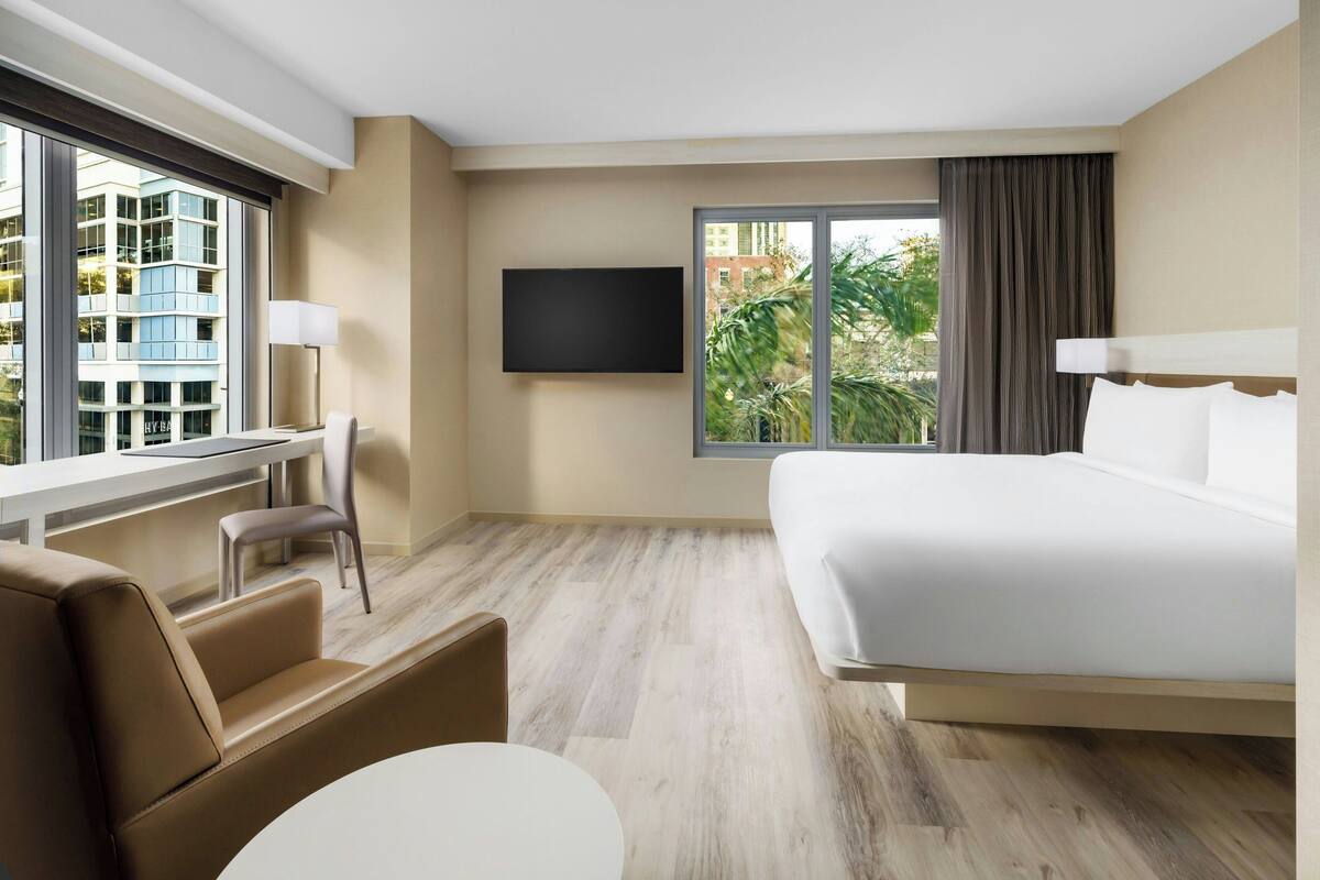 A modern hotel room with a large bed, a desk, and a chair. There is a TV mounted on the wall and a window with a view of buildings and trees.