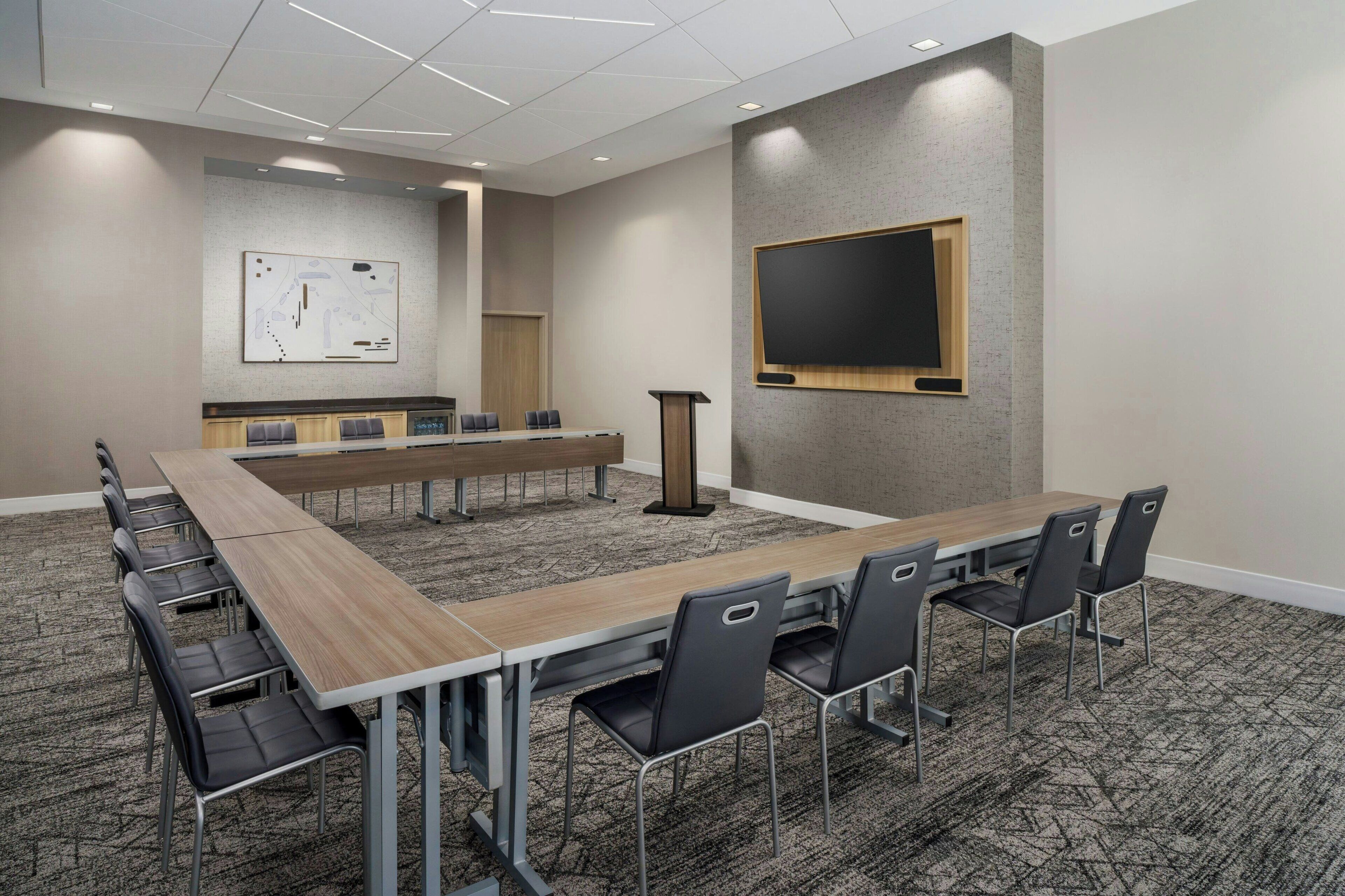 Une salle de conférence avec une longue table, des chaises noires, un pupitre et un tableau blanc au mur.