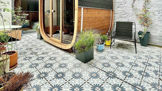 Un patio urbano moderno con puerta de cristal, plantas en macetas, un banco y baldosas estampadas.