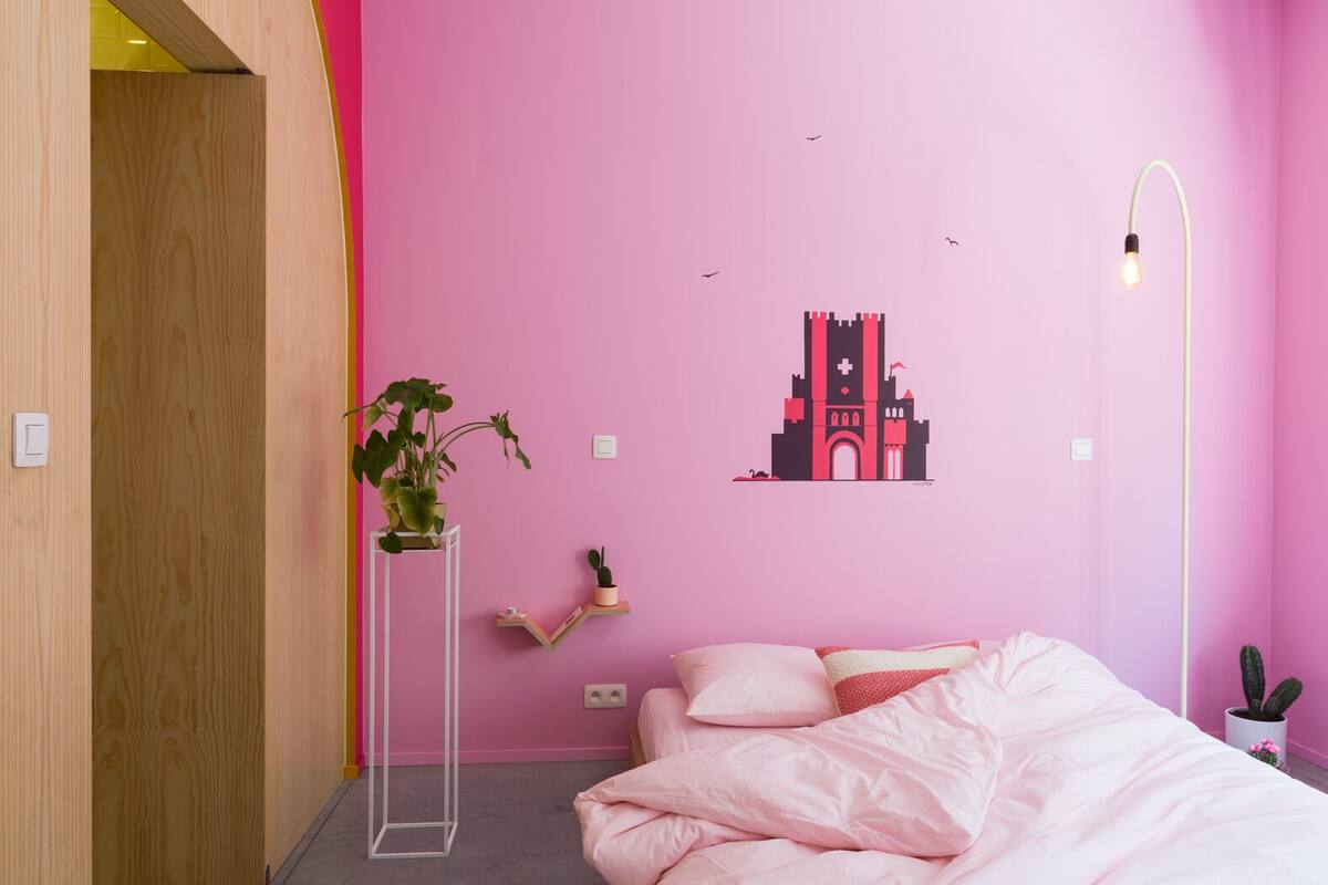Une chambre avec un mur rose, un lit avec une literie rose, une plante en pot, une étagère fixée au mur et une œuvre d’art représentant un bâtiment.