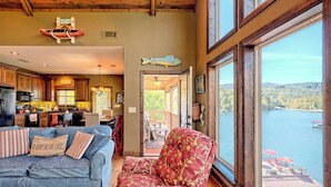 A spacious living area with a large window overlooking a lake, a cozy seating area with a patterned armchair, and a kitchen with wooden cabinets and a dining table.