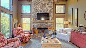 A cozy living room with a stone fireplace, a television mounted above it, and a comfortable seating area with a red patterned armchair and a white sofa.