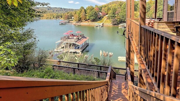 A wooden deck with a view of a lake, a dock with a boat, and a house in the background.