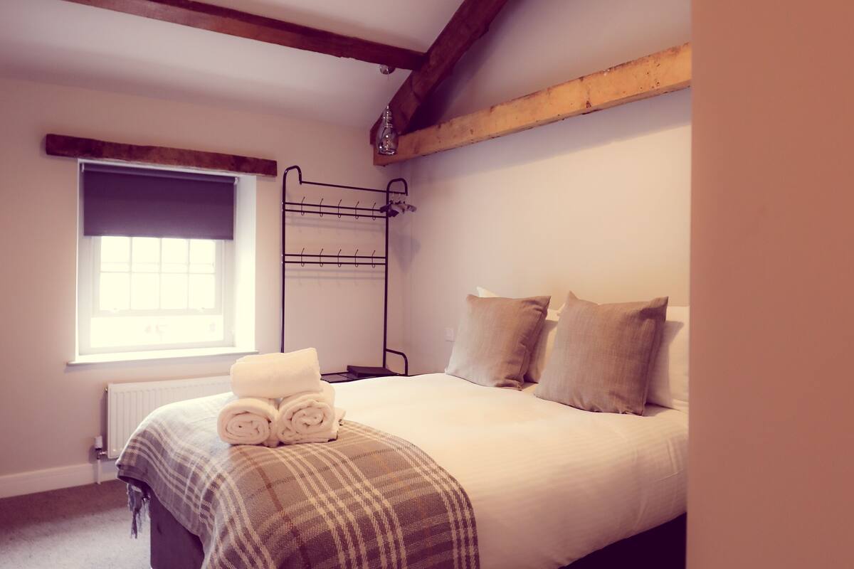 A bedroom with a bed, a window, a radiator, and a wooden ceiling beam.