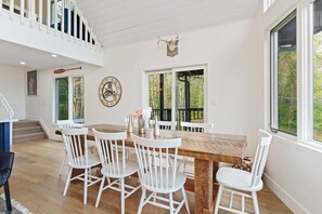 A dining area with a wooden table and white chairs, a clock, and a window with a view of trees.