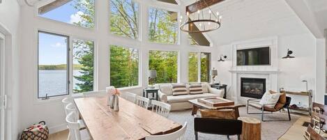 A spacious living area with a large wooden dining table, white chairs, a fireplace, and a view of the outdoors.