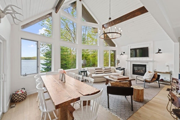 A spacious living area with a large wooden dining table, white chairs, a fireplace, and a view of the outdoors. - Muskokan Mirage Located on Mirage Lake (Novar)