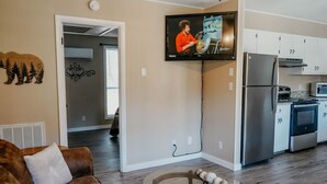 A modern living room with a brown leather sofa, a glass coffee table, and a flat-screen TV mounted on the wall. A kitchen with white cabinets and stainless steel appliances is visible in the background.