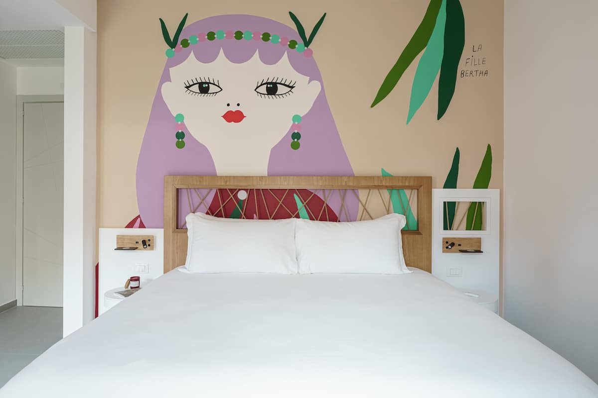 Un dormitorio con cama, cabecera de madera, mesitas de noche blancas y un mural en la pared.