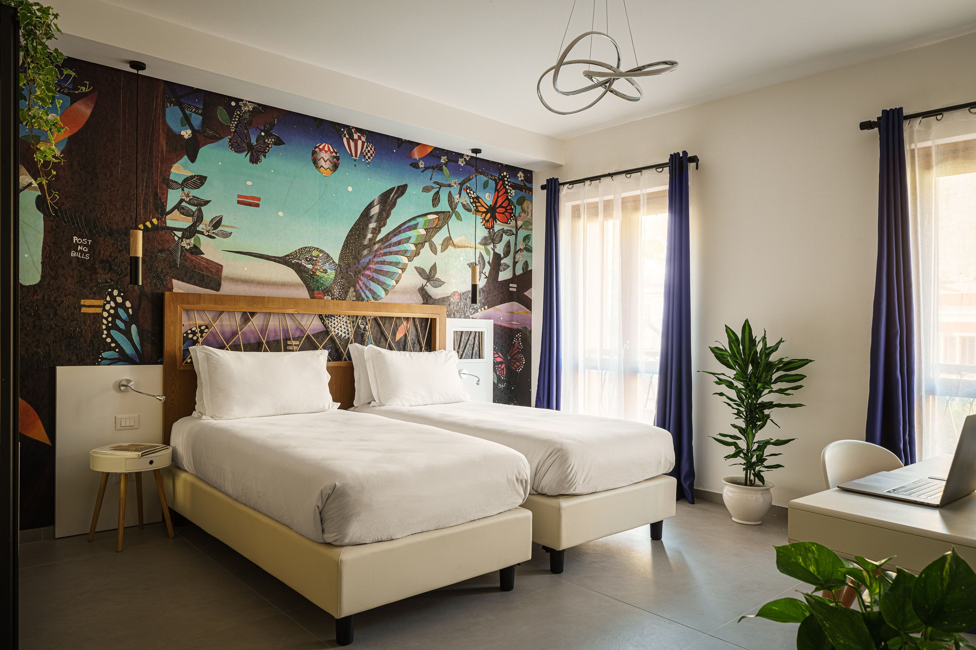 A hotel room with two beds, a desk with a laptop, a large butterfly mural on the wall, and a potted plant.