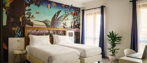 A hotel room with two beds, a desk with a laptop, a large butterfly mural on the wall, and a potted plant.